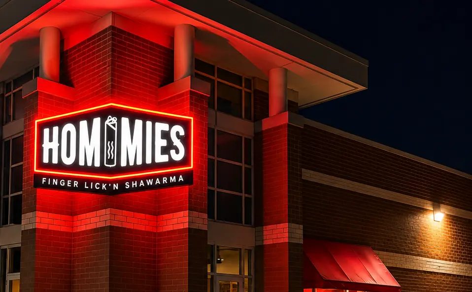 Hommies illuminated cabinet sign Hommies restaurant photographed at night on a brick corner unit: an illuminated cabinet sign reading HOMMIES above the line FINGER LICK'N SHAWARMA, mounted on the corner column under a canopy, with red accent lighting washing the brickwork and a red canopy to one side.